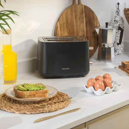 A matte black Salter two-slice toaster sits on a white kitchen countertop, styled with avocado toast on a plate, a carton of brown eggs, a moka pot, and wooden cutting boards in the background.