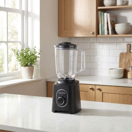 A black countertop blender with a clear pitcher sits on a white kitchen counter in a bright kitchen with wood cabinets and a white subway tile backsplash.