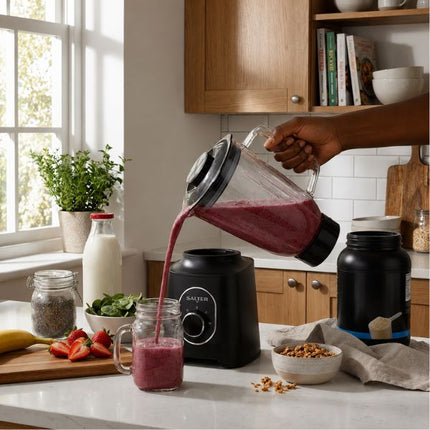 A hand pours a pink smoothie from a black blender pitcher into a glass jar on a kitchen counter, with a black blender base, a black supplement container, strawberries, oats, and other kitchen items in the background.