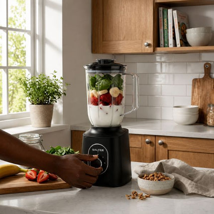 A black Salter blender filled with yogurt, strawberries, and greens on a kitchen counter, with a person's hand adjusting the dial. The kitchen has light wood cabinets, white subway tile, and a marble countertop.