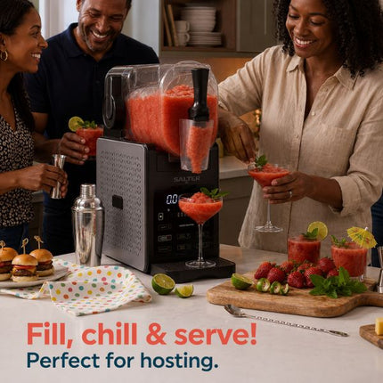 A Salter slushie maker on a kitchen counter with three people holding margarita glasses, surrounded by strawberries, limes, and drinks. The Salter brand is visible on the machine.
