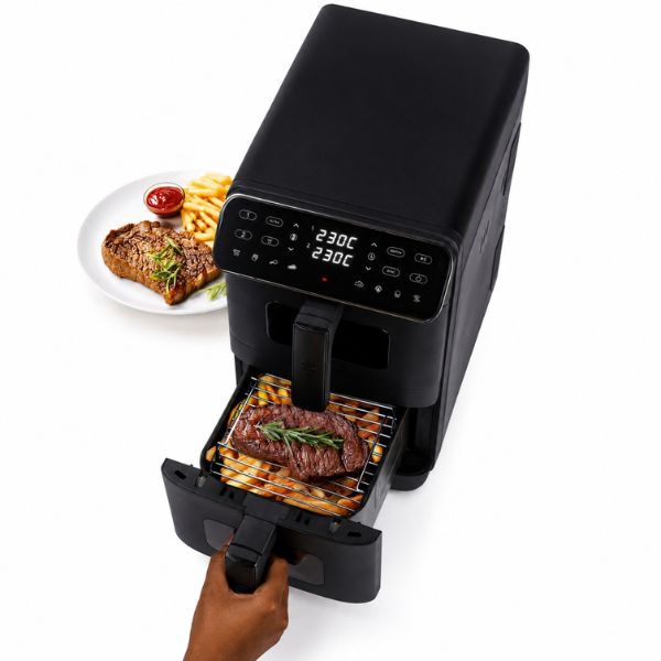 A black air fryer with its drawer pulled open, showing a steak cooking inside. A hand is holding the handle, and a plate of steak and fries sits to the left on a white background.