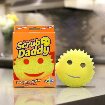A yellow smiley face textured scrubber next to its orange packaging box labeled 'The Original Scrub Daddy'.