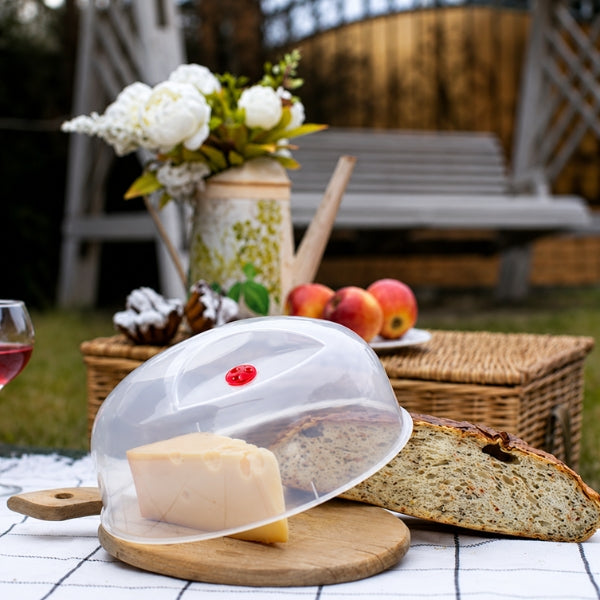 Sozali transparent microwave cover covering cheese on a wooden board beside sliced bread at an outdoor picnic.