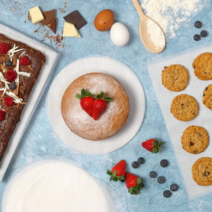 Overhead baking scene with a strawberry-topped round cake sitting on a Tala greaseproof circle, with cookies, brownies and a stack of Tala 23 cm greaseproof circles 