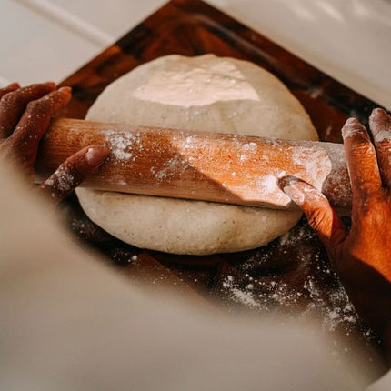 Hands rolling out a round ball of dough with a wooden rolling pin on a dark wooden board dusted with flour.
