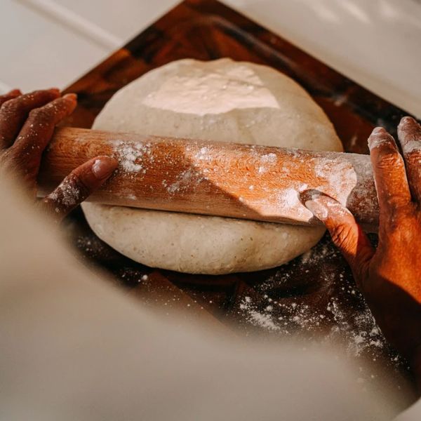 Hands rolling out a round ball of dough with a wooden rolling pin on a dark wooden board dusted with flour.