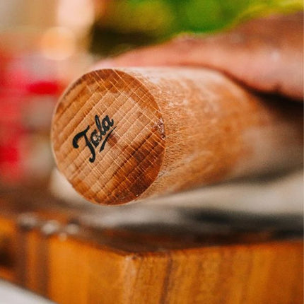 A close-up of a wooden rolling pin resting on a wooden cutting board, with a hand holding the handle. The end of the rolling pin is engraved with the brand name 'Tola' in black script.