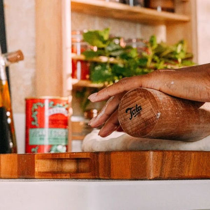A wooden rolling pin with the brand name 'Tala' engraved on its end, being used by a person's hand to roll out dough on a wooden board in a warm kitchen setting.