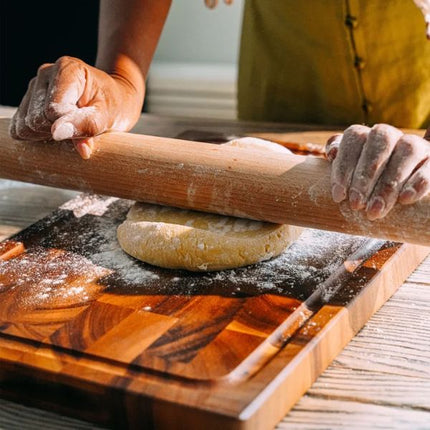 A close-up lifestyle shot of a person using a wooden rolling pin to flatten dough on a wooden cutting board with flour dusting.