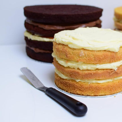 Tala small angled palette knife beside a layered vanilla cake topped with white frosting, with a chocolate cake behind
