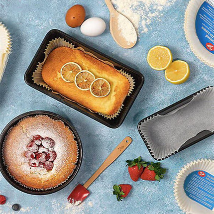 A top-down styled baking scene on a light blue surface showing two baked goods: a rectangular loaf cake with lemon slices in a black loaf pan and a round cake topped with berries in a round pan. A second empty black loaf pan with parchment paper, lemons, strawberries, eggs, a wooden spoon, flour, and baking paper are arranged around it.