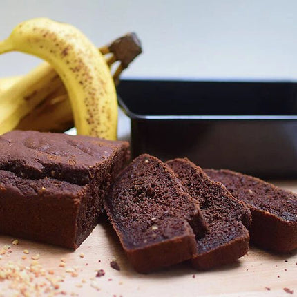 Slices of chocolate banana bread on a wooden surface with whole bananas and a black loaf pan in the background.