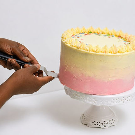 Hands using a Tala angled spreader spatula to smooth pink buttercream on a yellow-and-pink frosted cake whilst being placed on a cake stand