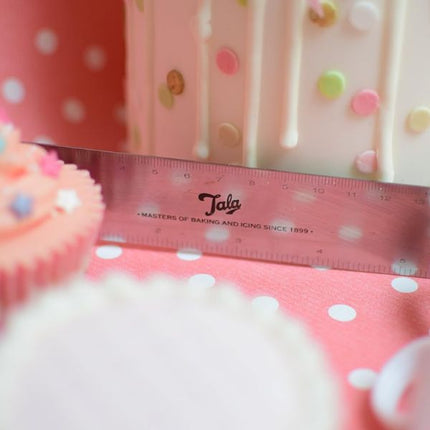 Close-up of a Tala angled spreader spatula with measurement markings beside a pastel drip cake with sugar decorations