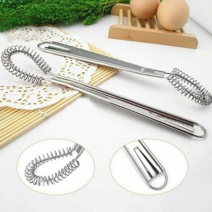 A stainless steel kitchen utensil with a long handle and spiral spring end, shown on a white surface with decorative props including bamboo sticks, a lace doily, greenery, eggs, and a wooden egg stand. Two circular inset close-ups at the bottom show detail views of the spring and handle.